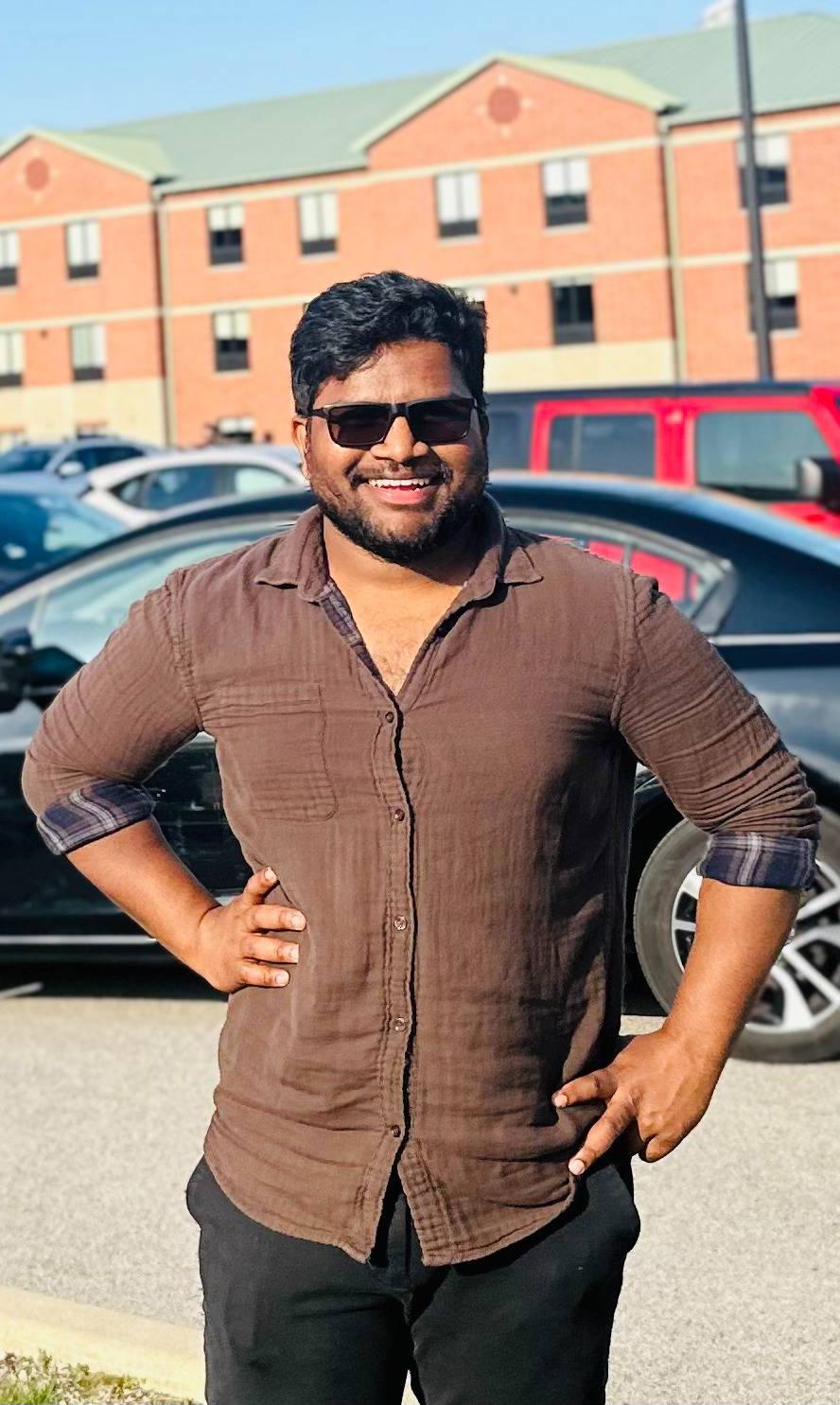 Student in brown shirt and sunglasses smiling at the camera with brick building in the background